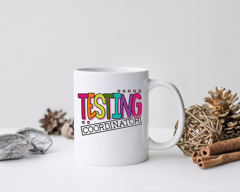 Op de afbeelding: Witte keramische mok met de tekst "TESTING COORDINATOR" in kleurrijke, dikgedrukte letters. De mok heeft een gebogen handvat en staat tegen een neutrale achtergrond met decoratieve elementen.