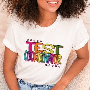 May include: White t-shirt with the text "TEST COORDINATOR" in colorful letters. The letters are outlined in black and have a dotted pattern around them.