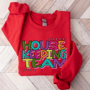 May include: Red sweatshirt with the text "HOUSE KEEPING TEAM" in colourful letters.