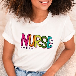 Peut inclure: T-shirt blanc avec le mot "NURSE" en lettres color&eacute;es et soulign&eacute;es. Les lettres sont dans un arc-en-ciel de couleurs avec du rose, de l'orange, du jaune, du vert et du bleu.