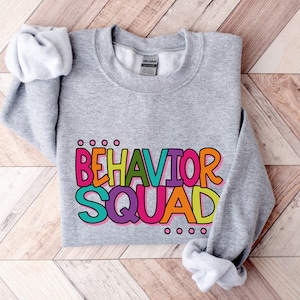 May include: Gray sweatshirt with the text "BEHAVIOR SQUAD" in colorful, outlined letters. The letters are in different colors, including pink, yellow, green, blue, and orange.