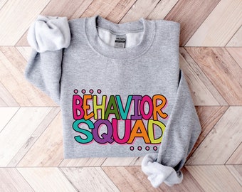 Könnte beinhalten: Grauer Pullover mit dem Text "BEHAVIOR SQUAD" in bunten, umrandeten Buchstaben. Die Buchstaben sind in verschiedenen Farben, darunter Pink, Gelb, Grün, Blau und Orange.