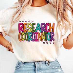 May include: Cream-colored t-shirt with the words "RESEARCH COORDINATOR" in a colorful, bubble-style font. The text is outlined in black and features pink, orange, green, and purple hues. The shirt is worn with blue jeans.