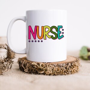 Peut inclure: Une tasse en c&eacute;ramique blanche avec un graphisme color&eacute; qui dit "NURSE:" avec quatre points violets en dessous du texte.