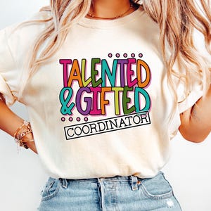 May include: A cream-colored t-shirt featuring a multi-colored graphic print on the chest. The text reads TALENTED & GIFTED in bold, colorful letters, with the word COORDINATOR printed in black inside a rectangular box underneath the main text.