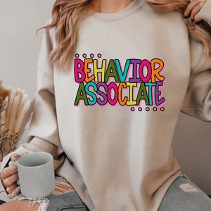 May include: A light gray sweatshirt with the text "BEHAVIOR ASSOCIATE" in colorful letters. The letters are outlined in black and have a pink, yellow, green, blue, and orange color scheme.