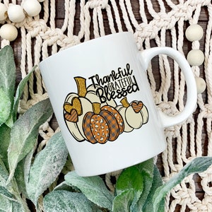 May include: White ceramic coffee mug with a handle. The mug features a design of three pumpkins with gold glitter accents and the text "Thankful Grateful Blessed".