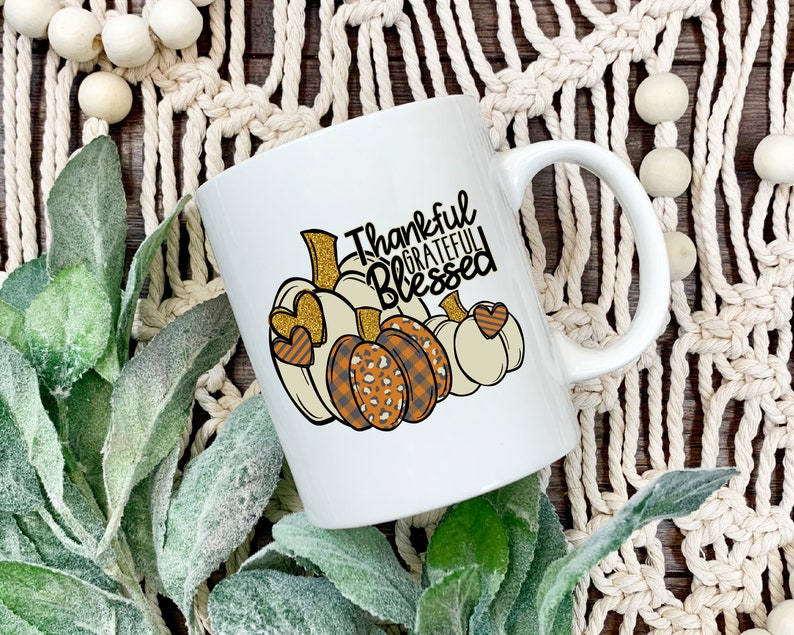 May include: White ceramic coffee mug with a handle. The mug features a design of three pumpkins with gold glitter accents and the text "Thankful Grateful Blessed".