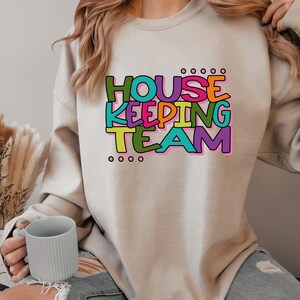 May include: A light beige sweatshirt with the text "HOUSE KEEPING TEAM" in colourful letters. The letters are outlined in black and have a playful, hand-drawn style.