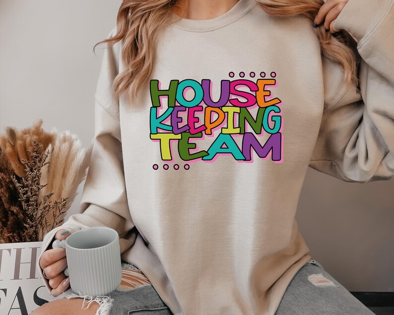 May include: A light beige sweatshirt with the text "HOUSE KEEPING TEAM" in colourful letters. The letters are outlined in black and have a playful, hand-drawn style.