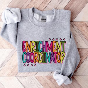 May include: A heather gray sweatshirt with the words "ENRICHMENT COORDINATOR" in a colorful, block letter design. The text is outlined in black and filled with shades of pink, orange, yellow, green, and blue. The sweatshirt has long sleeves and a crew neck.