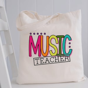 以下が含まれることがあります： レインボーフォントで「MUSIC TEACHER」と書かれたカラフルなグラフィックが施された白いキャンバス地のトートバッグです。