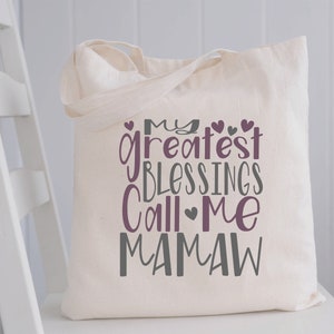 Peut inclure: Un sac fourre-tout en toile blanche avec le texte "My greatest blessings call me Mamaw" en lettres violettes et grises. Le texte est entour&eacute; de petits c&oelig;urs.