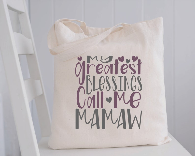 Peut inclure: Un sac fourre-tout en toile blanche avec le texte "My greatest blessings call me Mamaw" en lettres violettes et grises. Le texte est entour&eacute; de petits c&oelig;urs.