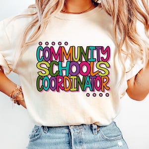 May include: A cream-colored short-sleeve t-shirt featuring the text "COMMUNITY SCHOOLS COORDINATOR" printed in a multi-colored, bold, rounded font. The letters are outlined in black and filled with various colors including pink, green, orange, and teal.