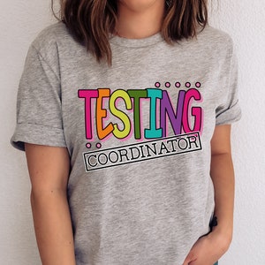 Op de afbeelding: Een grijs t-shirt met de tekst "TESTING COORDINATOR" in een kleurrijk, omlijnd lettertype. De tekst is roze, oranje, groen en paars. Het shirt heeft opgerolde mouwen.