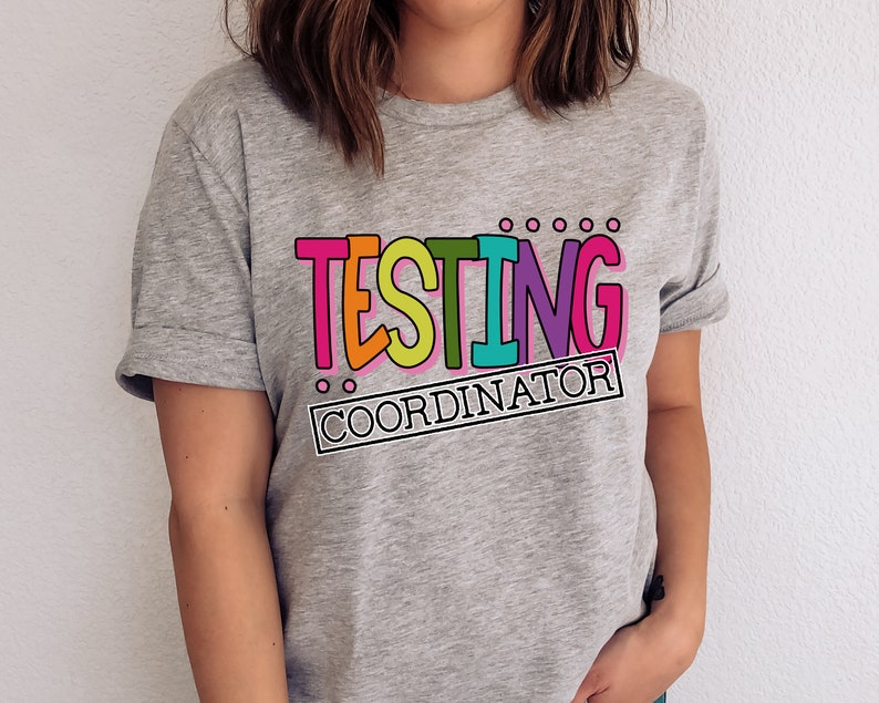 Op de afbeelding: Een grijs t-shirt met de tekst "TESTING COORDINATOR" in een kleurrijk, omlijnd lettertype. De tekst is roze, oranje, groen en paars. Het shirt heeft opgerolde mouwen.