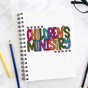 May include: A white notebook with a colorful handwritten title "CHILDREN'S MINISTRY" on the front cover. The notebook is open and has a spiral binding. There are three pencils and a pair of glasses on a white surface next to the notebook.
