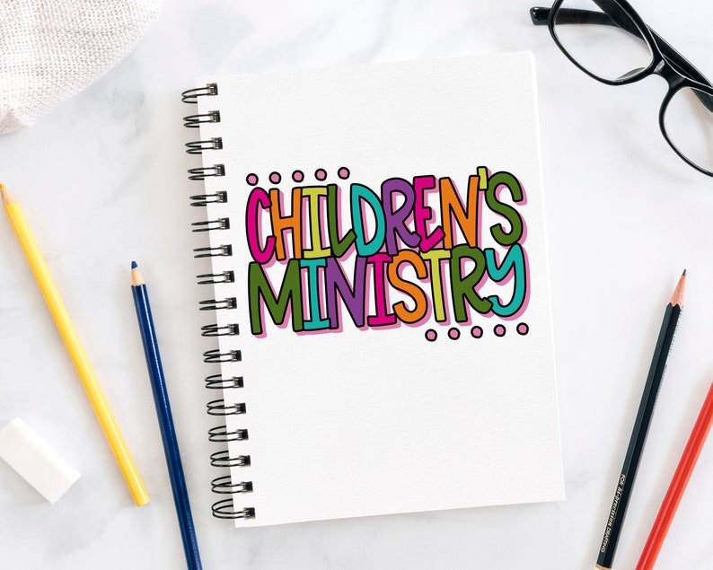 May include: A white notebook with a colorful handwritten title "CHILDREN'S MINISTRY" on the front cover. The notebook is open and has a spiral binding. There are three pencils and a pair of glasses on a white surface next to the notebook.