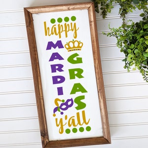May include: A wooden sign with the text "happy Mardi Gras y'all" in gold, green, and purple lettering. The sign is decorated with a crown, a jester's hat, and a mask.