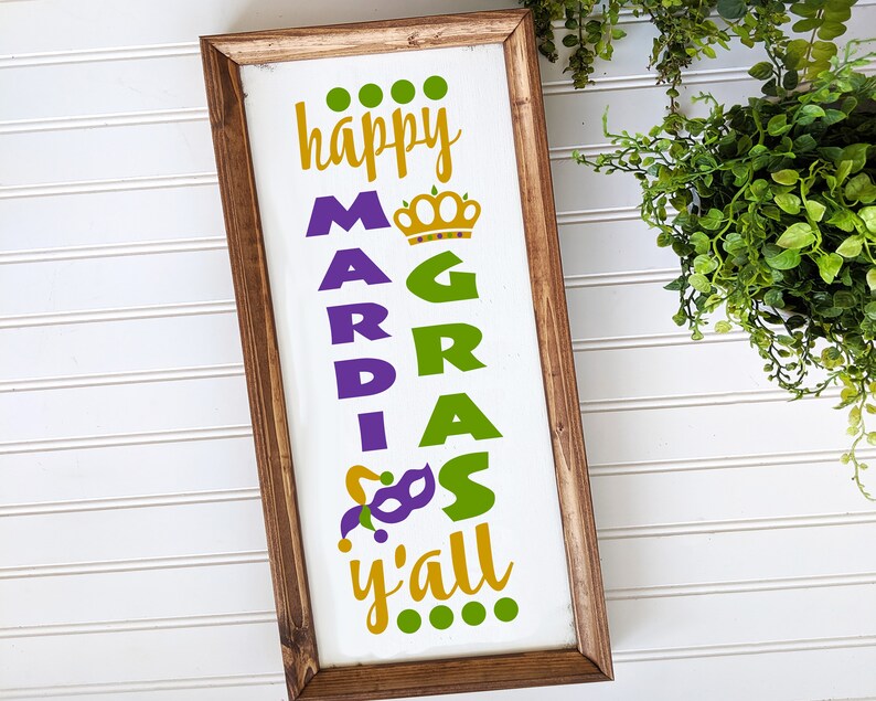 May include: A wooden sign with the text "happy Mardi Gras y'all" in gold, green, and purple lettering. The sign is decorated with a crown, a jester's hat, and a mask.