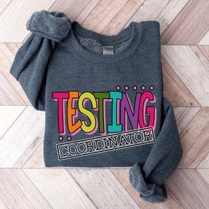 Op de afbeelding: Een donkergrijze sweatshirt met de tekst "TESTING COORDINATOR" in een kleurrijk, vetgedrukt lettertype. De tekst is zwart omlijnd en staat tegen een achtergrond van roze, oranje, groen, paars en blauw. De sweatshirt ligt op een houten oppervlak.