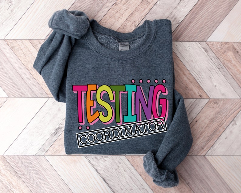 Op de afbeelding: Een donkergrijze sweatshirt met de tekst "TESTING COORDINATOR" in een kleurrijk, vetgedrukt lettertype. De tekst is zwart omlijnd en staat tegen een achtergrond van roze, oranje, groen, paars en blauw. De sweatshirt ligt op een houten oppervlak.