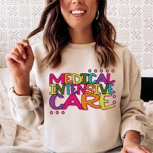 May include: A beige sweatshirt with the words "MEDICAL INTENSIVE CARE" in a colorful, bubble letter design. The text is outlined in black and features shades of pink, green, orange, and purple. The sweatshirt is a neutral color.