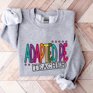 May include: Gray sweatshirt with the text "ADAPTED PE TEACHER" in colorful letters.