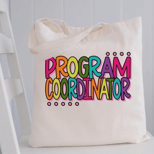May include: A cream-colored tote bag with the words "PROGRAM COORDINATOR" in large, colorful letters. The text is outlined in black and features a rainbow of colors, including pink, orange, green, teal, purple, and yellow. The bag has two handles.