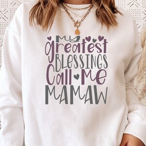 Peut inclure: Sweat-shirt blanc avec le texte "My greatest blessings call me Mamaw" en lettres violettes et grises avec de petits c&oelig;urs.