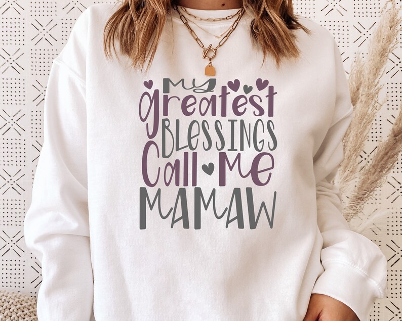 Peut inclure: Sweat-shirt blanc avec le texte "My greatest blessings call me Mamaw" en lettres violettes et grises avec de petits c&oelig;urs.