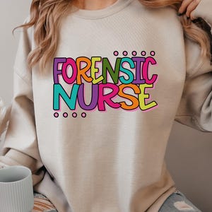 May include: A beige sweatshirt with the words "FORENSIC NURSE" in colorful, bubble-style letters. The text is outlined in black and features a variety of colors, including purple, orange, green, and yellow. The sweatshirt is a neutral color.