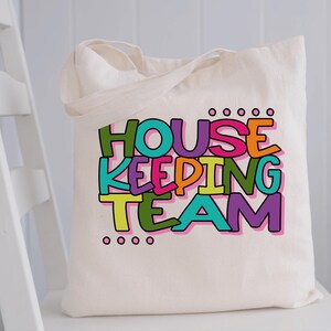 May include: A white canvas tote bag featuring the text "HOUSEKEEPING TEAM" in colourful letters. The letters are outlined in black and have a playful, hand-drawn style.