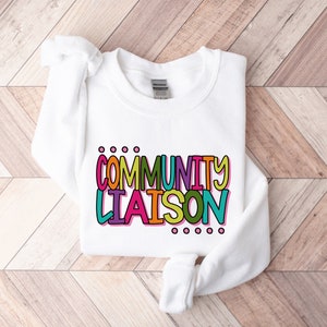 May include: White sweatshirt with the text "COMMUNITY LIAISON" in colorful, outlined letters.