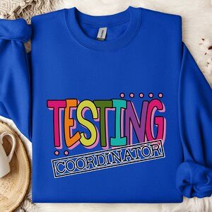 Op de afbeelding: Een koningsblauwe sweatshirt met de tekst "TESTING COORDINATOR" in een kleurrijk, retro lettertype. De tekst is zwart omlijnd en geplaatst tegen een witte achtergrond. De sweatshirt is gemaakt van een zacht materiaal.