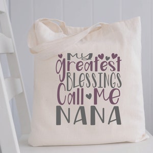 May include: A white canvas tote bag with the text "My greatest blessings call me Nana" in purple and gray lettering. The text is stylized with hearts.