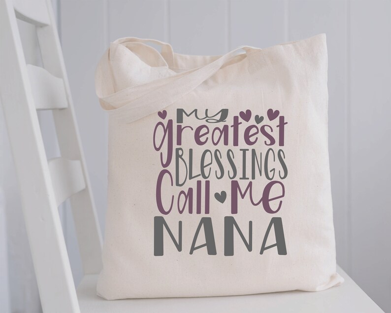 May include: A white canvas tote bag with the text "My greatest blessings call me Nana" in purple and gray lettering. The text is stylized with hearts.
