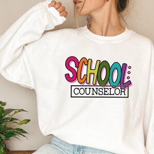 以下が含まれることがあります： レインボーカラーの「SCHOOL: COUNSELOR」のグラフィックがプリントされた白いスウェットシャツです。