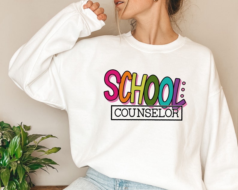 以下が含まれることがあります： レインボーカラーの「SCHOOL: COUNSELOR」のグラフィックがプリントされた白いスウェットシャツです。