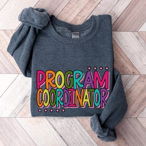 May include: A dark gray sweatshirt with the words "PROGRAM COORDINATOR" in colorful bubble letters. The text is outlined in black and has a playful, cartoonish style. The sleeves are folded.