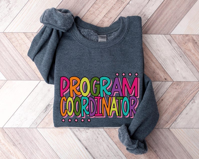 May include: A dark gray sweatshirt with the words "PROGRAM COORDINATOR" in colorful bubble letters. The text is outlined in black and has a playful, cartoonish style. The sleeves are folded.