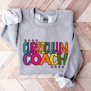 May include: Gray sweatshirt with the text "CURRICULUM COACH" in colorful letters. The letters are outlined in black and have a rainbow color scheme.