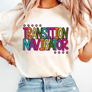 May include: A cream-colored short-sleeve t-shirt featuring the text "TRANSITION NAVIGATOR" printed in a bold, multi-colored font with shades of pink, orange, yellow, green, and teal, accented by small circles above and below the words.