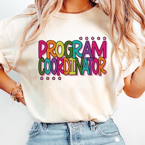 May include: Cream-colored t-shirt with the words "PROGRAM COORDINATOR" in a colorful, outlined font. The text is in pink, orange, green, and blue. The shirt is worn with blue jeans.