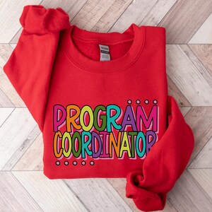 May include: A red sweatshirt with the words "PROGRAM COORDINATOR" in colorful, bubble letters. The text is outlined in black and has small dot accents above and below the words. The sweatshirt is folded, with the sleeves visible.
