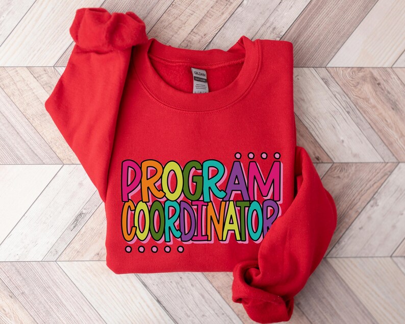 May include: A red sweatshirt with the words "PROGRAM COORDINATOR" in colorful, bubble letters. The text is outlined in black and has small dot accents above and below the words. The sweatshirt is folded, with the sleeves visible.