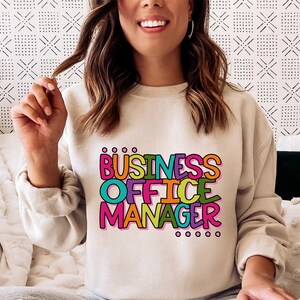 以下が含まれることがあります： 明るいベージュのトレーナーに、カラフルな輪郭のある文字で「BUSINESS OFFICE MANAGER」と書いてあります。テキストは遊び心のある漫画のフォントです。