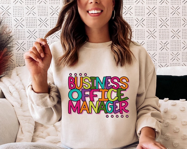 以下が含まれることがあります： 明るいベージュのトレーナーに、カラフルな輪郭のある文字で「BUSINESS OFFICE MANAGER」と書いてあります。テキストは遊び心のある漫画のフォントです。