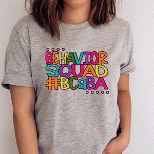May include: Gray t-shirt with a colorful graphic that says "Behavior Squad #BCaBA".
